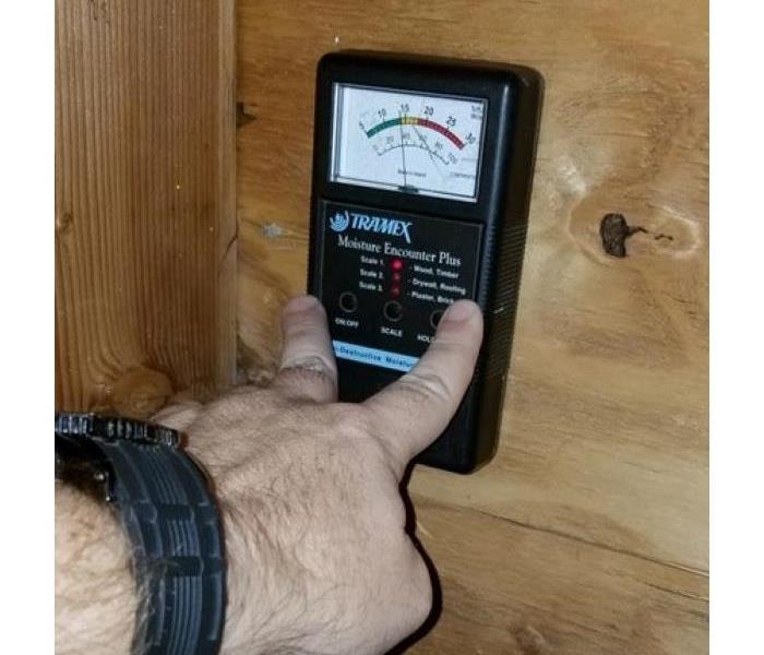 Photo is showing a man's hand holding a moisture meter down on a wood subfloor to get moisture readings