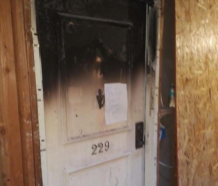Photo is showing a burned and charred front door of an apartment after fire has damaged it.